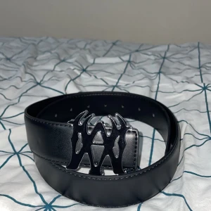 Svart bälte med unik spänne - Snyggt svart bälte med ett iögonfallande metallspänne i silver. Perfekt för att ge din outfit en edgy touch. Bältet har flera hål för justering och är tillverkat i ett robust material. Jättefin bälte och är helt ny, vill sälja den för att jag har flera bälten och jag behöver inte den