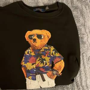 Säljer en svart tröja från Ralph Lauren med det ikoniska Polo Bear-motivet. Björnen har en färgglad hawaiiskjorta och solglasögon, vilket ger en avslappnad och cool look. Perfekt för dig som gillar stil med en twist! Pris kan diskuteras vid snabb affär.