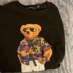 Svart tröja med Polo Bear från Ralph Lauren - Säljer en svart tröja från Ralph Lauren med det ikoniska Polo Bear-motivet. Björnen har en färgglad hawaiiskjorta och solglasögon, vilket ger en avslappnad och cool look. Perfekt för dig som gillar stil med en twist! Pris kan diskuteras vid snabb affär.