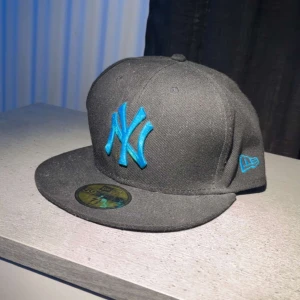 Svart keps från New Era  - Snygg svart keps från New Era med blå broderad logga framtill. Kepsen har en klassisk design med platt skärm och är perfekt för en casual look. Den är tillverkad i ett slitstarkt material och har en bekväm passform. Perfekt sick typ ny har använt den typ 4 gånger  pris ej hugget i sten.