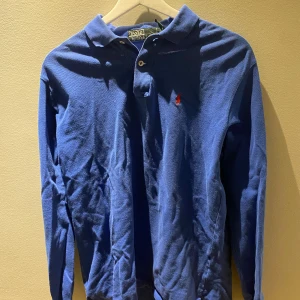 Blå långärmad pikétröja från Ralph Lauren - Säljer en klassisk blå långärmad pikétröja från Ralph Lauren. Tröjan har en liten röd logga på bröstet och en krage med knappar. Perfekt för en stilren och avslappnad look. Xl i barn så passar M