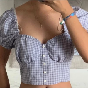 Sommartopp - Säljer en söt lila rutig topp från H&M Divided med korta puffärmar och knappar framtill. Toppen är croppad och har en smickrande passform. Perfekt för en somrig look!