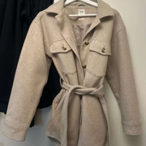 Beige kappa från Lee - Snygg beige kappa från Lee med knappar och bälte i midjan. Jackan har långa ärmar och två stora fickor framtill. Perfekt för höst och vår!