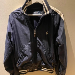 Marinblå vårjacka från Ralph lauren  - Snygg svart bomberjacka från Polo Ralph Lauren med vita ränder på kragen och ärmarna. Jackan har dragkedja och två fickor framtill. Perfekt för en stilren look.