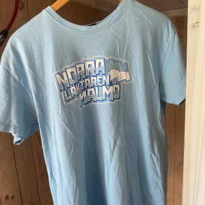 Ljusblå t-shirt norra läktaren Malmö - Säljer en ljusblå t-shirt med tryck från Norra Läktaren Malmö. T-shirten har en normal passform och är kortärmad, perfekt för en avslappnad stil. Materialet är mjukt och bekvämt.