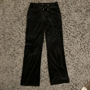 Svarta velourbyxor Juicy Couture - Svarta mjukisbyxor från Juicy Couture i glansig velour med dragsko i midjan och fickor både fram och bak. Byxorna har raka ben och snygga detaljer med knappar på bakfickorna. Perfekta för en chill och trendig look.