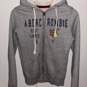 Grå hoodie från Abercrombie & Fitch - Säljer en grå hoodie från Abercrombie & Fitch med dragkedja och broderad logga framtill. Tröjan har en justerbar huva med snören och två fickor. I storlek S. 