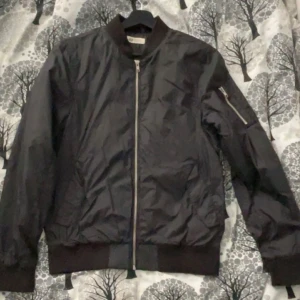 Svart bomberjacka med dragkedja - Snygg svart bomberjacka med dragkedja framtill och en ficka med dragkedja på ärmen. Jackan har ribbade muddar vid ärmslut och nederkant för en klassisk look. Perfekt för en stilren och avslappnad stil.