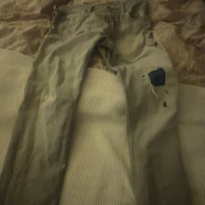 Gråa jeans med slitningar - Säljer ett par gröna jeans med coola slitningar och en lappad detalj på ena benet. Perfekta för en avslappnad stil. De har en klassisk femficksdesign och knappgylf.