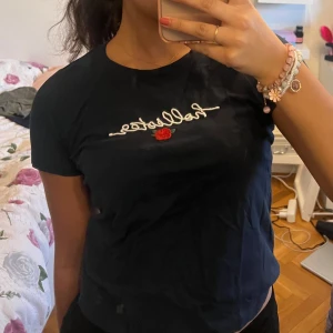 Mörkblå t-shirt från Hollister - Säljer en mörkblå t-shirt från Hollister med broderad text och en liten röd ros på framsidan. T-shirten har en klassisk rund hals och korta ärmar. Perfekt för en avslappnad stil.