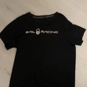 Svart t-shirt från Sail Racing - Snygg svart t-shirt från Sail Racing med vit logga på bröstet och en mindre logga på ryggen. Perfekt för en avslappnad stil.