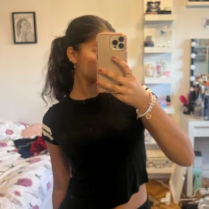 Svart t-shirt med vita ränder - Säljer en svart t-shirt med vita ränder på ärmarna. Den har en croppad passform och korta ärmar. Perfekt för en avslappnad stil.