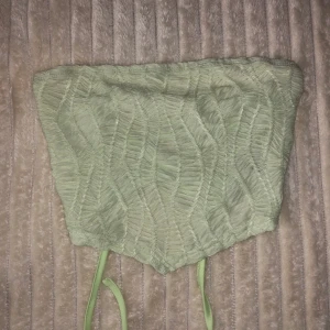 Grön korsettopp med snörning - Snygg grön korsettopp med ett unikt mönster och snörning i ryggen. Perfekt för att ge en stilfull touch till din outfit. Toppen är axelbandslös och har en croppad design. Använd 2ggr. Det är i storlek S men passar även M