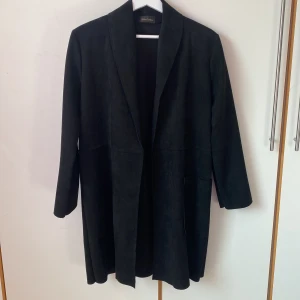 Svart längre kavaj från Chica London - Elegant svart kavaj från Chica London. Öppen och rak modell utan knappar eller dragkedja. Tillverkad i ett mjukt mockaliknande material. Stora rymliga fickor. Längd 90 cm, axelbredd 43. Saknar storleksmärkning men är ca M/L. 