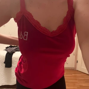 Röd linne med spetsdetaljer - Snygg röd linne med spetsdetaljer och strassbokstäver 'B&B' på bröstet. Tillverkad i ett sammet material som ger en lyxig känsla. Perfekt för en kväll ute eller en speciell tillställning.