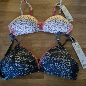 Två nya mönstrade BH:ar från Esprit 70AA - Säljer två snygga BH:ar från Esprit. De är helt nya med etiketterna kvar. Den ena är vit med blommigt mönster och rosa detaljer, medan den andra är svart med stjärnor och blommor. Båda har justerbara axelband och en bekväm passform. Nypris 398.