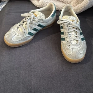 Gazelle Adidas sneakers - Gazelle Adidas sneakers med klassiska gröna ränder på sidan. Skorna har en gummisula och snörning framtill. Perfekta för en avslappnad stil. Det finns en defekt, skorna har tvättats vilket ledde till att den gröna färgningen fick hela vita skon att bli grå/beigeaktig som det syns på bilderna. 