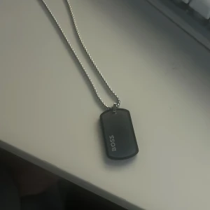 Svart halsband med bricka från Boss - Stilrent halsband med en svart bricka från Boss. Brickan hänger på en silverfärgad kedja och har en modern och minimalistisk design. Perfekt för att ge en elegant touch till din outfit.