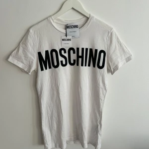 Vit t-shirt från Moschino - Säljer en stilren vit t-shirt från Moschino med stort svart tryck på framsidan. Perfekt för en casual look med en touch av lyx. T-shirten är kortärmad och gjord i mjukt bomullsmaterial. Allt original är med!