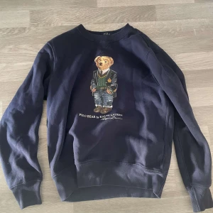 Mörkblå sweatshirt från Polo Ralph Lauren - Säljer en mörkblå sweatshirt från Polo Ralph Lauren med en ikonisk Polo Bear-design på framsidan. Tröjan har långa ärmar och en rund halsringning. Perfekt för en avslappnad stil. Mycket fint skick, hör av er vid eventuella frågor eller funderingar! 
