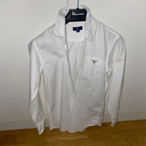 Vit skjorta från GANT - Klassisk vit skjorta från GANT med broderad logga på bröstfickan. Skjortan har långa ärmar och knappar framtill. Perfekt för en stilren look.