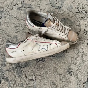 Vita sneakers från Golden Goose - Snygga vita sneakers från Golden Goose med silverdetaljer och en stjärna på sidan. Skorna har en cool, sliten look och snörning framtill. Perfekta för en avslappnad stil med en touch av lyx. Äktighetsbevis finns