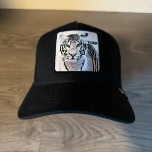 Svart keps med tigerbroderi från Goorin Bros - Snygg svart keps från Goorin Bros med ett coolt broderi av en tiger på framsidan. Perfekt för att ge din outfit en djärv touch. Kepsen har en justerbar passform och är en del av The Farm-kollektionen.