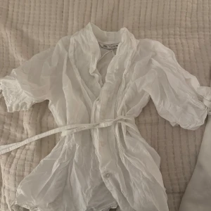 Vit blus från Zara - Säljer en elegant vit blus från Zara med knytband i midjan. Blusen har en luftig design med trekvartsärmar och knappar framtill. Perfekt för en stilren look.💕