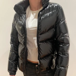 Svart glansig dunjacka från Colmar - Snygg svart glansig dunjacka från Colmar med dragkedja och hög krage. Jackan har en pufferdesign och är perfekt för kyligare väder. Den har långa ärmar och en stilren look med Colmar-logga på ärmen. Stl 14, ca 162 cm.