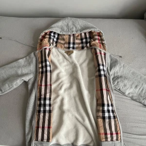 Grå hoodiejacka från Burberry XL - Snygg grå hoodiejacka från Burberry i bomull med dragkedja och klassiskt rutigt foder i huvan. Jackan har snörning i huvan och ribbade muddar. Perfekt för dig som vill ha en stilren och bekväm look med lyxig känsla.