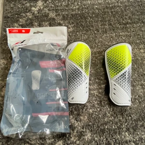 Pro Touch benskydd S vit/gul - Säljer ett par oanvända benskydd från Pro Touch i storlek S. De är vita med coola gula och svarta detaljer och har justerbara svarta kardborreband på baksidan. Skydden är tillverkade i polypropylen, polyester, TPU och gummi för bästa komfort och skydd på fotbollsplanen. Pris kan diskuteras. 