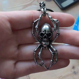 Halsband med spindel och dödskalle - Coolt halsband med svart snöre och en stor silverfärgad spindelberlock som har en dödskalle som kropp. Perfekt accessoar för dig som gillar alternativ stil eller vill sticka ut med något unikt och edgy. 