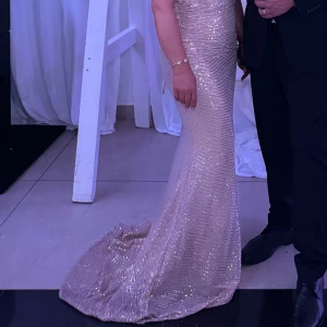 Champagnefärgad glittrig långklänning - Säljer en elegant långklänning i champagnefärg med glittriga paljetter och fjäderdetaljer vid bysten. Klänningen är figurnära och axelbandslös, perfekt för dig som vill sticka ut på bal eller fest. Materialet är glittrigt och skimrande, vilket ger en lyxig känsla.