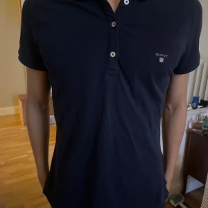 Mörkblå pikétröja från Gant - Snygg mörkblå pikétröja från Gant med klassisk krage och två knappar framtill. Tröjan har korta ärmar och diskret Gant-logga broderad på bröstet. Perfekt för en clean och stilren look.