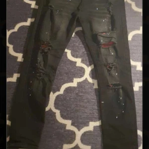 Svarta skinny jeans med slitningar - Svarta skinny jeans med flera slitningar och hål framtill, samt stänk av vit färg för en edgy look. Jeansen har smal passform och är tillverkade i stretchigt denimtyg. Perfekta för dig som gillar streetstyle och vill sticka ut.