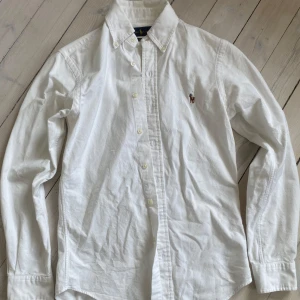 Vit skjorta Ralph Lauren Slim Fit XS - Vit skjorta från Ralph Lauren i slim fit-modell med klassisk button down-krage och broderad logga på bröstet. Tillverkad i  bomull. Långärmad och med knappar framtill. Perfekt för en clean och stilren look.