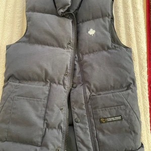 Mörkgrå dunväst från Canada Goose - Säljer en mörkgrå dunväst från Canada Goose med klassisk quiltad design och två stora fickor framtill. Västen har dragkedja och tryckknappar, samt en broderad lönnlöv-detalj på bröstet. Yttermaterialet är i polyester och bomull, och fyllningen är av dun.