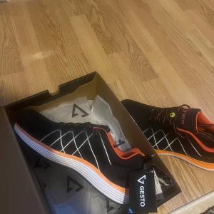 Svarta sneakers från Gesto med orange detaljer - Snygga svarta sneakers från Gesto med orange insida och detaljer samt vita grafiska linjer på sidorna. Skorna har snörning och en vit och orange sula som ger en sportig vibe. Perfekta för dig som gillar att sticka ut med färg och stil. Splitter nya aldrig använda med lappar kvar 