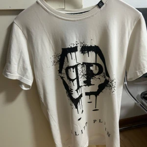 Vit Philipp Plein t-shirt med tryck storlek M - Snygg vit t-shirt från Philipp Plein med stort svart grafiskt tryck framtill. Klassisk rund hals och korta ärmar. Skön bomullskvalitet som känns mjuk mot huden. Perfekt för dig som gillar streetstyle och vill sticka ut med en cool logga.