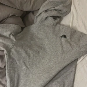 Grå hoodie från The North Face - Säljer en grå hoodie från The North Face med svart broderad logga på bröstet. Tröjan har huva med snörning och en klassisk känguruficka framtill. Perfekt för chill dagar eller när du vill ha en enkel och stilren look.
