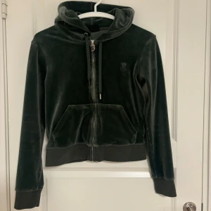 Mörkgrön velour hoodie Juicy Couture - Mörkgrön hoodie från Juicy Couture i mjuk velour med dragkedja framtill, snörning i huvan och känguruficka. Klassisk logga broderad på bröstet och ribbade muddar vid ärmslut och midja. Perfekt för en chill och trendig look.Varan är väl omhändertagen, men små defekter förekommer. Pris kan diskuteras, köparen står för frakt.