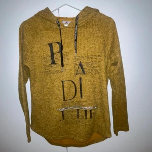 Hoodie - Gulmelerad hoodie med svart texttryck och coola bokstäver framtill. Tröjan har huva med svarta snören och långa ärmar. Mjuk och skön, perfekt för chill dagar.