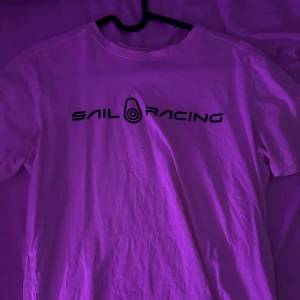Vit t-shirt från Sail Racing - Vit t-shirt från Sail Racing med svart logga tryckt över bröstet. Klassisk rund hals och korta ärmar. Perfekt basic-plagg med sportig vibe och stilren design.
