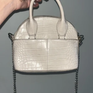 Beige handväska i crocopräglat från Zara - Snygg beige handväska från Zara med crocopräglat mönster. Väskan har två handtag, silverfärgad kedjerem och dragkedja upptill. Perfekt storlek för det viktigaste och ger en trendig touch till din outfit.