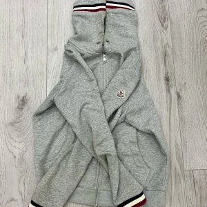Grå Moncler hoodie med dragkedja - Snygg grå hoodie från Moncler med hel dragkedja och klassisk logga på ärmen. Har tagit av snöret men kan skickas med om man vill! Ribbade muddar med röd, vit och marinblå rand. Perfekt för en chill och sportig look. Pris kan diskuteras