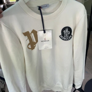 Vit Moncler sweatshirt med brodyr - Snygg vit sweatshirt från Moncler med rund halsringning. Tröjan har en stor broderad Moncler-logga på bröstet och ett guldigt broderat P. Mjuk bomullskvalitet och långa ärmar. Perfekt för dig som gillar stilrena och exklusiva plagg.