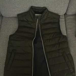 Grön dunväst med ståkrage (Strömberg) - Säljer en grön dunväst med ståkrage och horisontella quiltade sömmar. Västen har dragkedja framtill och två fickor på sidorna. På ryggen sitter en liten detalj. Perfekt för lager-på-lager-stil under kyliga dagar.