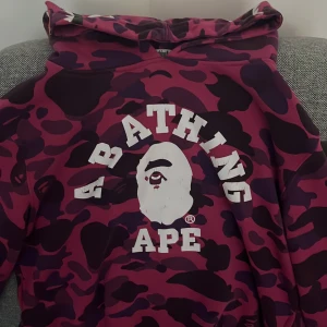 BAPE camo hoodie med shark-hood - Säljer en ikonisk BAPE hoodie i lila och vinröd camo med stor vit logga på bröstet. Huvan har shark-motiv, tigrer och broderade bokstäver. Perfekt statement-plagg för streetwear. 