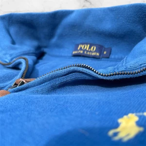 Blå half zip tröja från Polo Ralph Lauren - Väldigt bra skick förutom lite missfärgning vid sömarna, syns typ inte och är ganska snygga. Blå långärmad tröja från Polo Ralph Lauren med gul broderad logga på bröstet. Tröjan har en klassisk half zip-dragkedja med läderdetalj och är gjord i mjuk bomull. Perfekt för dig som gillar stilren och sportig look.