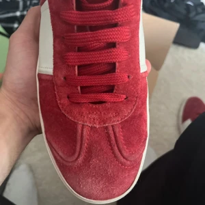 Valentino  röda sneakers mocka - Snygga Valentino Rockstud sneakers i röd mocka med vita detaljer och klassiska nitar på hälen. Skorna har rund tå, platt sula och röda snören. Perfekta för dig som vill sticka ut med en lyxig och trendig look. Lite slitna där fram och bak som man ser på bilderna 